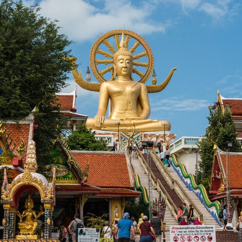 A golden statue of a large Buddha sitting on a pedestal, surrounded by smaller statues and colorful flags.