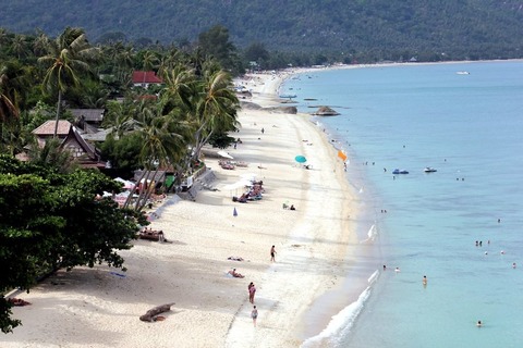 A scenic view of Lamai Beach on Koh Samui island in Thailand, with clear blue water, white sand, palm trees, and a boat in the distance.